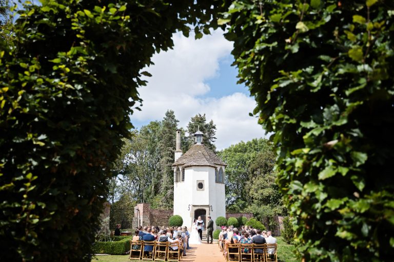 View through a hedge of a small castle and a bride and groom standing and saying their vows at their wedding. Wedding guests are seated.