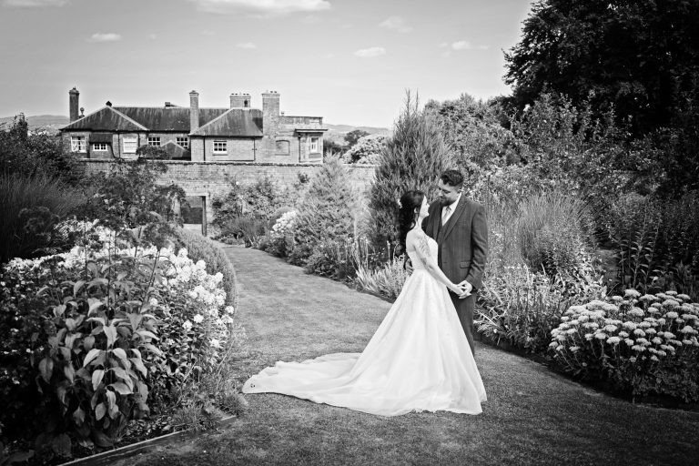 B&W image of a bride and groom holding hands romantically with a gorgeous Georgian house and gardens are behind them.