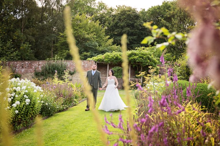 bride and groom hold hands whilst walking down an avenue of stunning plants inside a walled garden