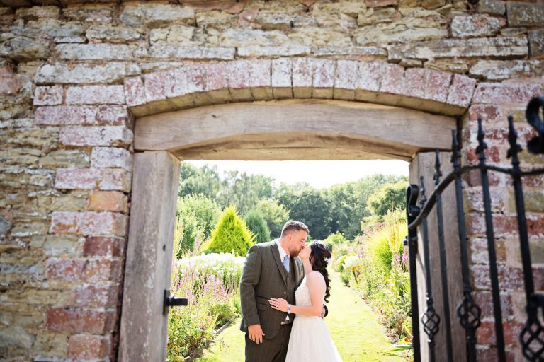 brick archway with a bride and groom kissing in the foreground. Behind them is a gorgeous kitchen garden