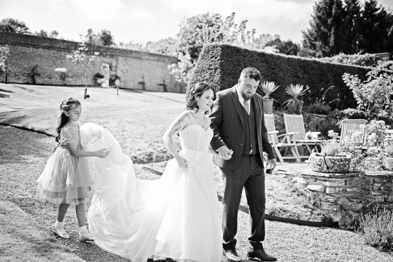 B&W image of a bride and groom walking holding hands whilst their daughter (flower girl) is carrying the train of her mums (bride) dress