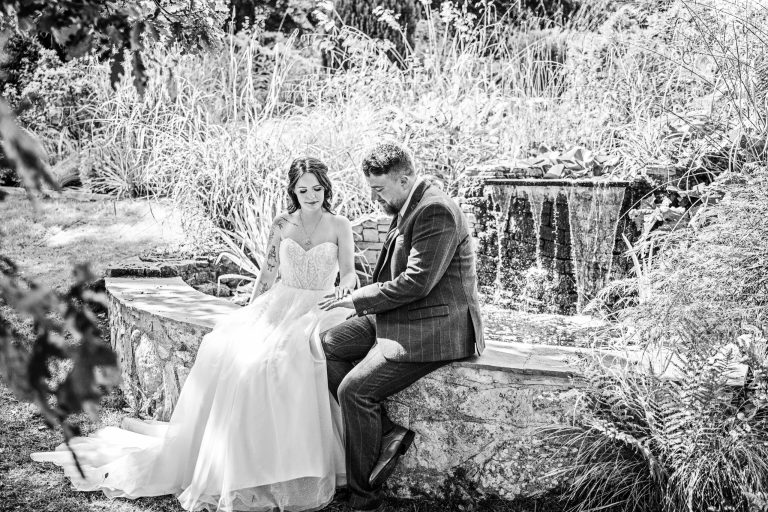 B&W image of a bride and groom, holding hands as they sit on a wall with a beautiful waterfall cascades behind them.