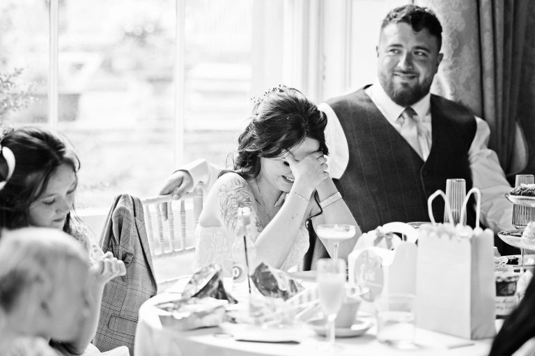 B&W photo of a bride surrounded by her groom and family with her hand on her head as she listens to a speech about her