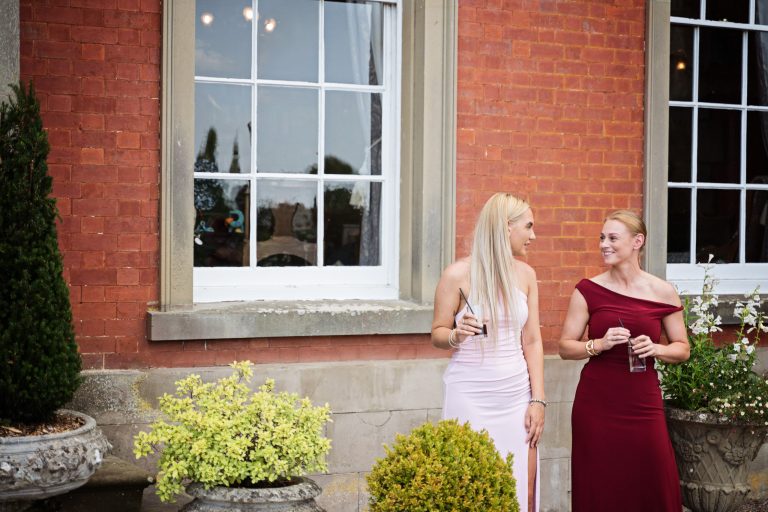 Two young ladies chat and smile at each other with a Georgian manor house wall and window behind them