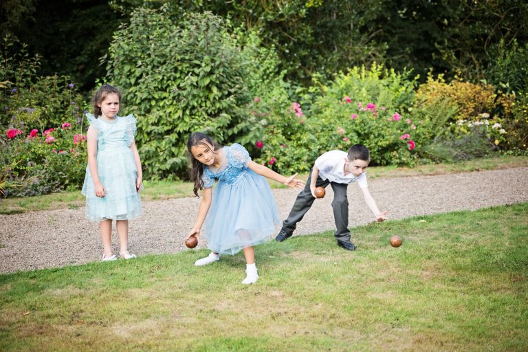 Three children at a wedding, one boy, one girl are throwing underarm patanque boules at the same time. The third child (girl) is watching them and where they throw.