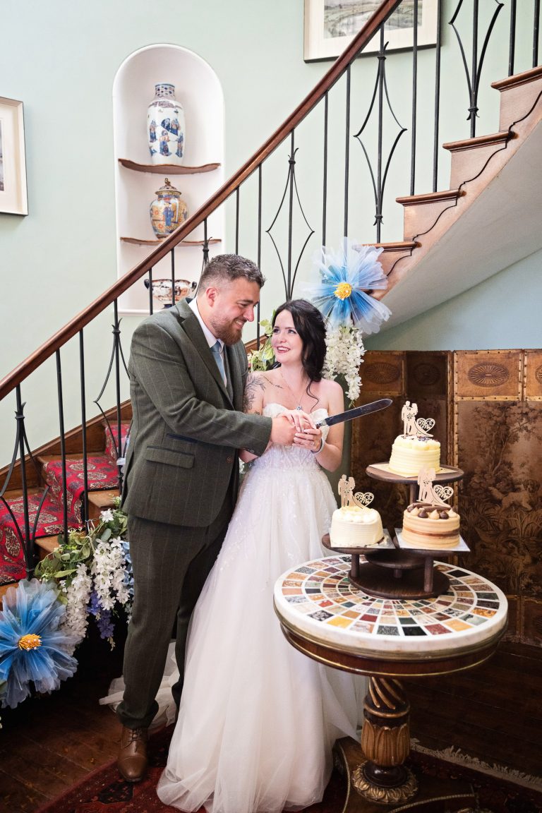 Bride and groom holding a cake knife as they are about to cut their wedding cake. A stunning georgian staircase slopes behind them.
