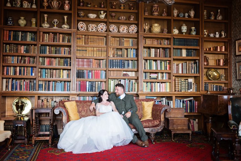 Library scene with a bride and groom sit on a chaise longue smiling at each other surrounded by books and a piano.