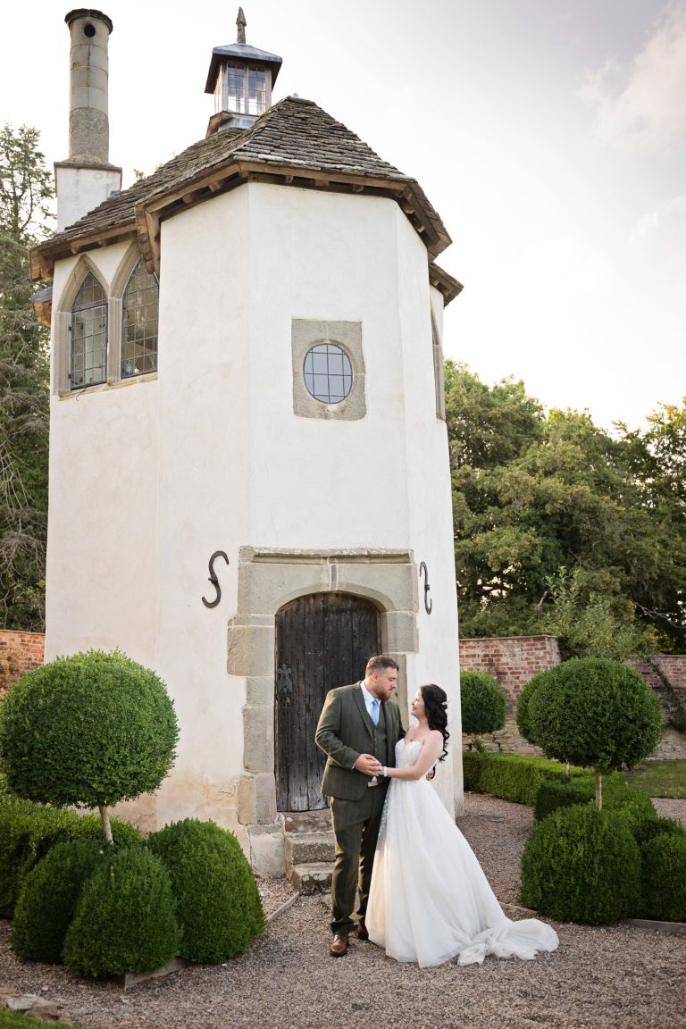 Portrait image of a bride and groom looking at each other and holding hands. A folly like tall building is behind them. Evening sunshine basks the couple.