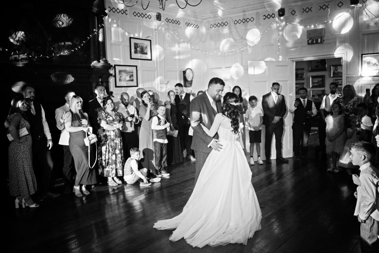 B&W image of a bride and groom doing their first dance with their friends and family watching